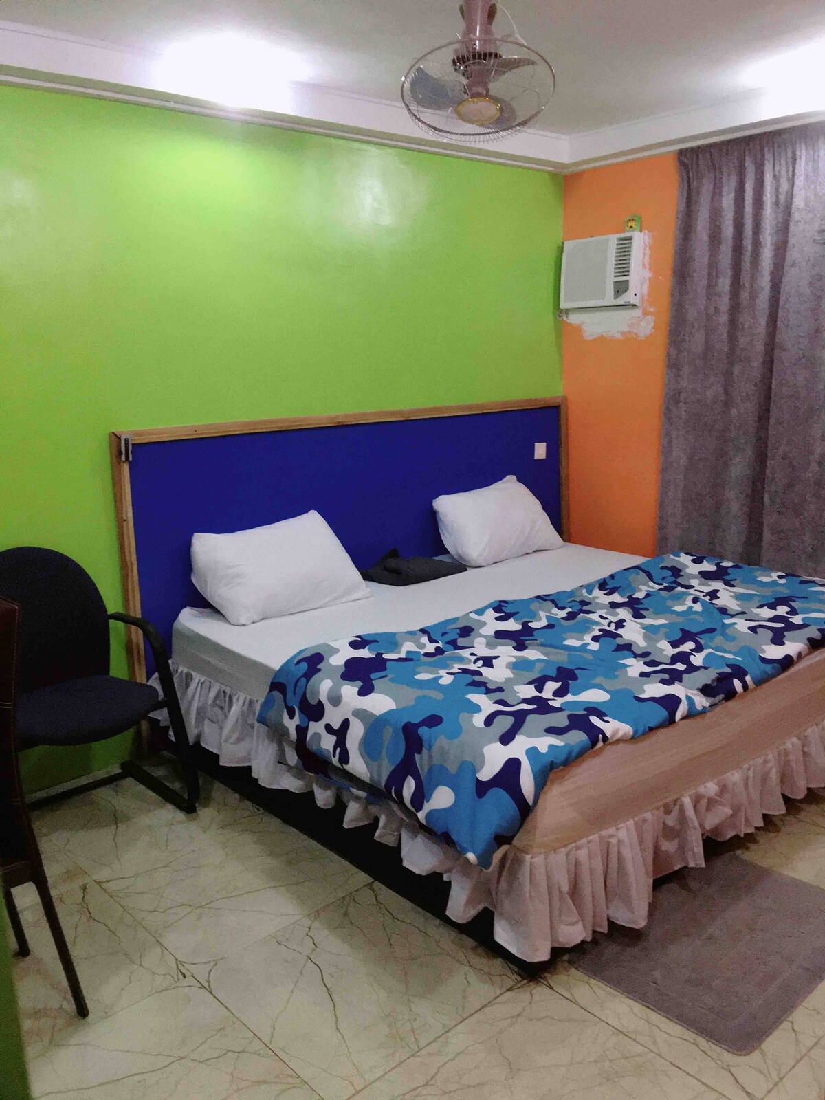 A comfortable room features a spacious bed adorned with a colorful blanket. A gray chair is positioned next to the bed. Green and orange walls create a lively atmosphere, with a window partially covered by dark curtains and a ceiling fan overhead. An air conditioning unit is also visible.