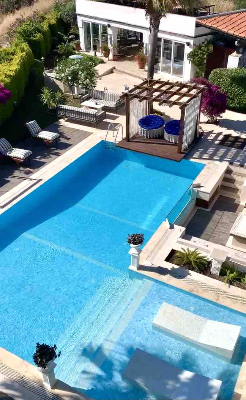 An aerial view showcases a serene pool area, featuring a turquoise water pool complemented by a wooden cabana adorned with cushions. Sun loungers are arranged beside the pool, with lush greenery and vibrant flowers enhancing the surroundings, creating a tranquil outdoor retreat.