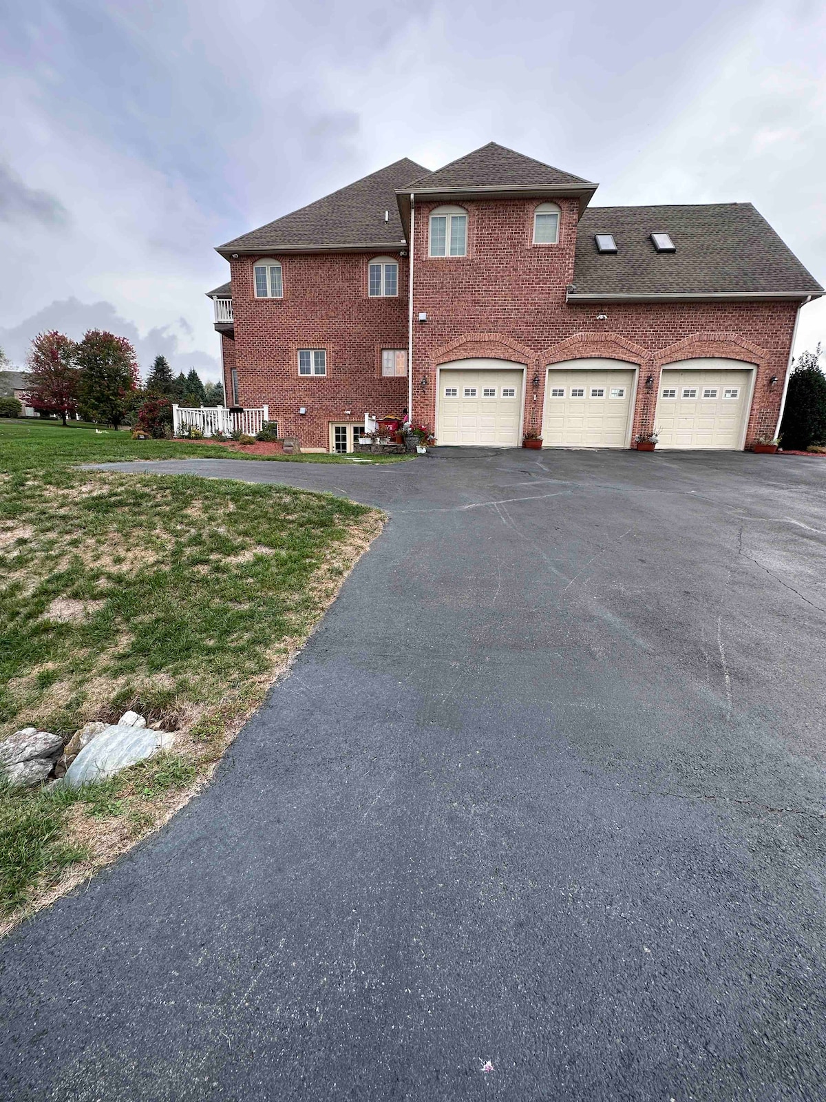 A large brick house is visible with three garage doors, set against a lush green lawn. The driveway leads up to the home, framed by trees and colorful plants. Soft clouds cover the sky, creating a serene backdrop.
