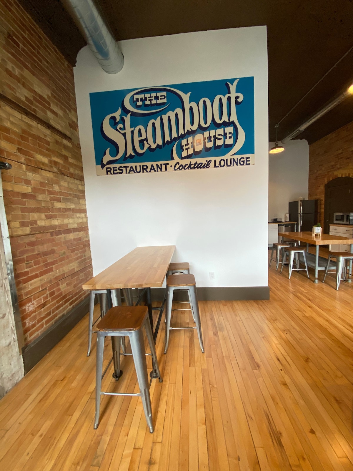 A rustic interior features exposed brick walls and polished wooden flooring. A large mural of 'The Steamboat House' is visible on one wall, complemented by high-top tables and metallic bar stools arranged nearby. The space is well-lit with overhead pendant lights.