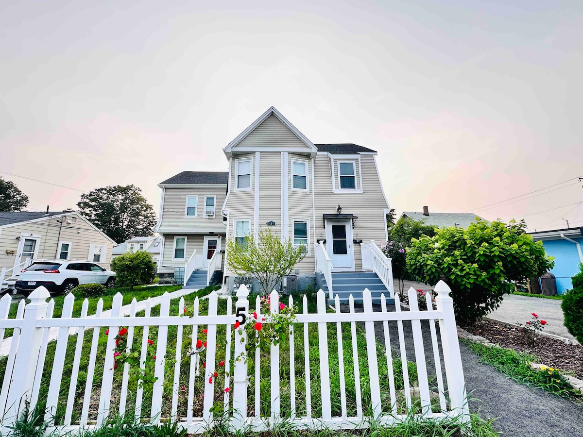 The multi-family house is set back from the street, featuring a neatly maintained yard bordered by a white picket fence. A pathway leads to the front entrance, highlighted by steps and surrounding greenery, with vehicles parked in the driveway.