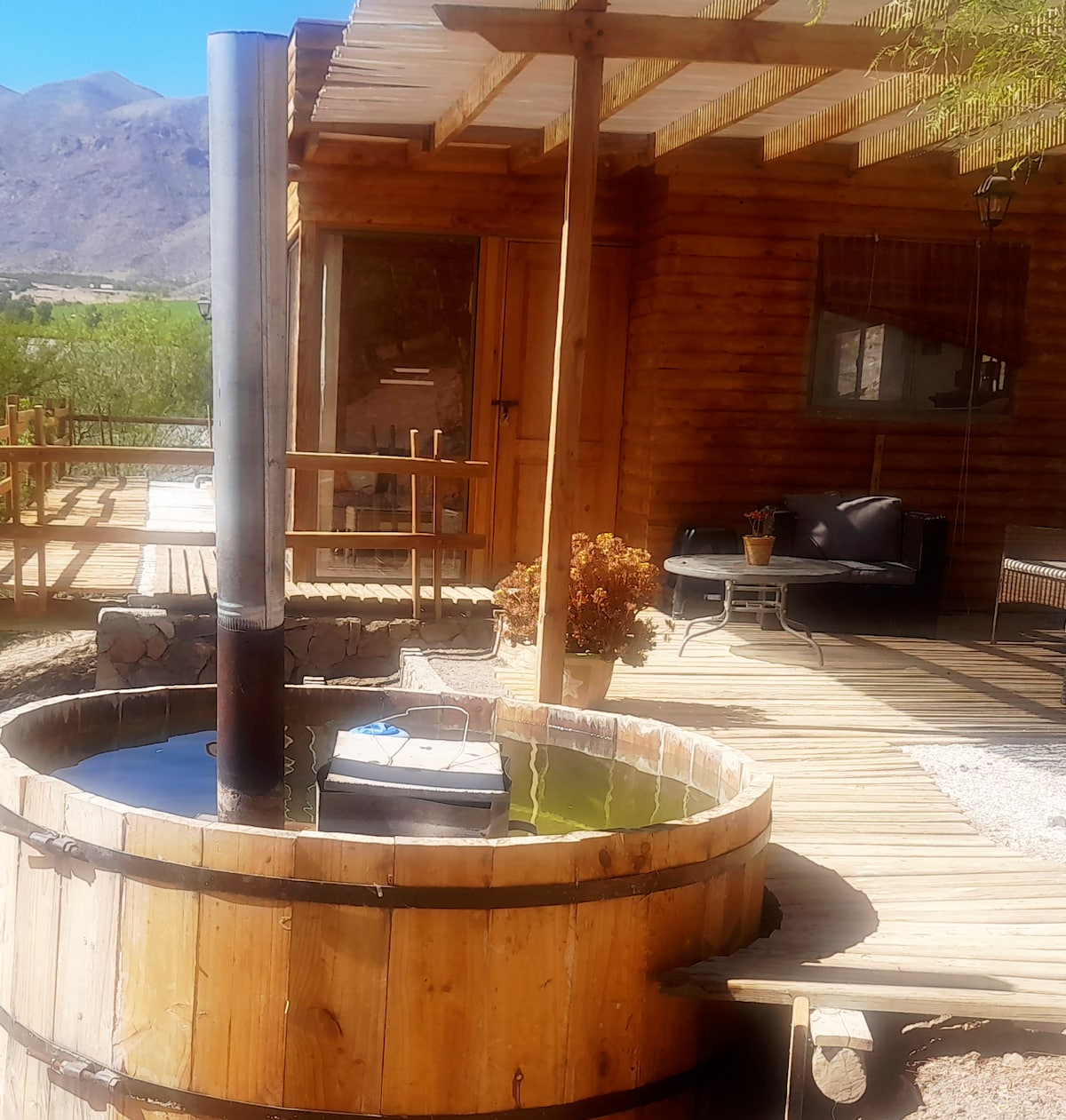 A rustic hot tub constructed from wood is positioned on a wooden deck, surrounded by outdoor seating. The natural scenery includes mountain views in the background, enhancing the serene environment. A small table with potted plants adds a touch of charm.