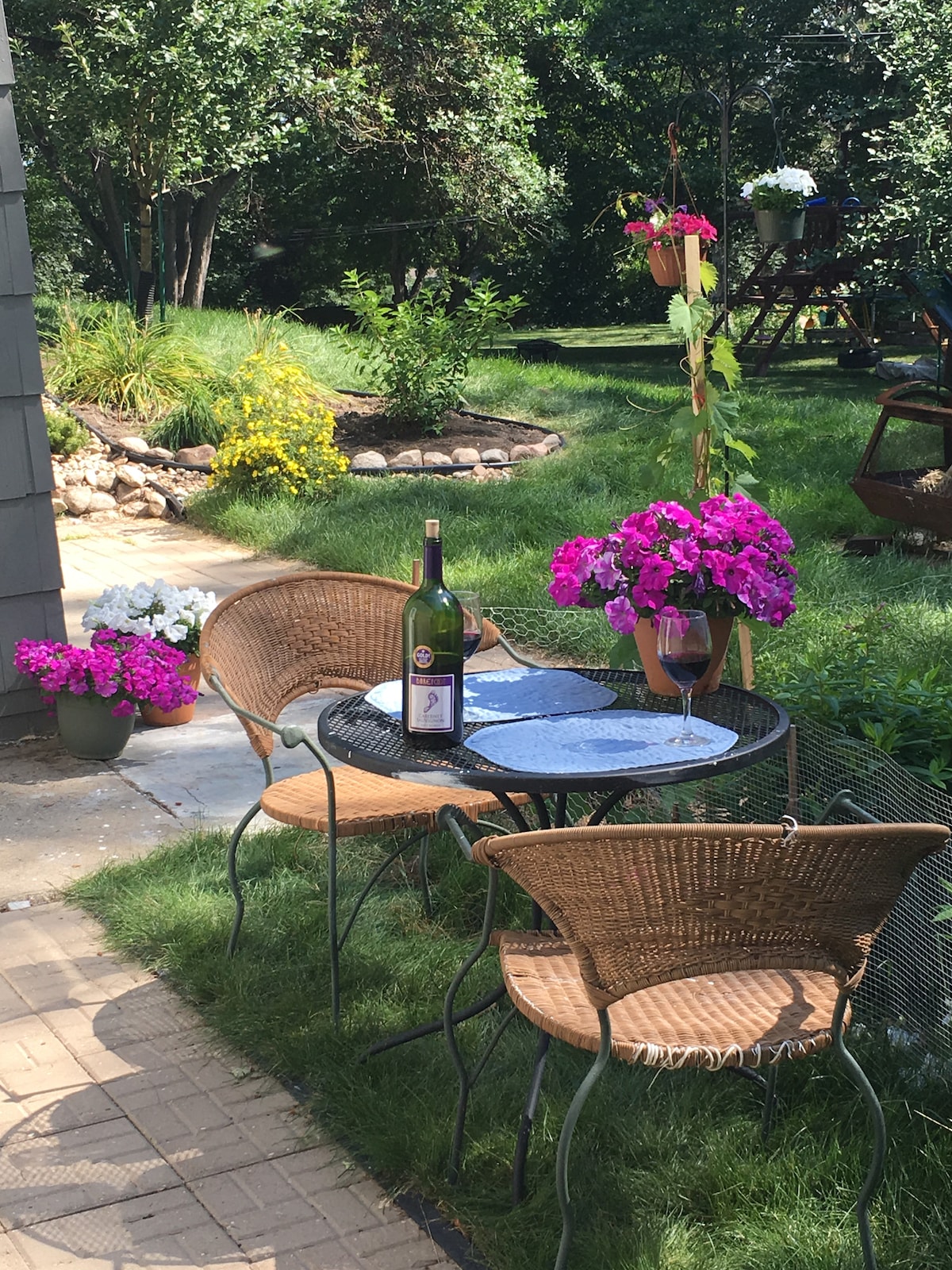 An outdoor patio set includes a round black table surrounded by two wicker chairs. A bottle of wine and a glass rest on the table, complementing vibrant pink and white flowers in nearby planters. A grassy area leads into a park-like setting with trees and colorful flora.