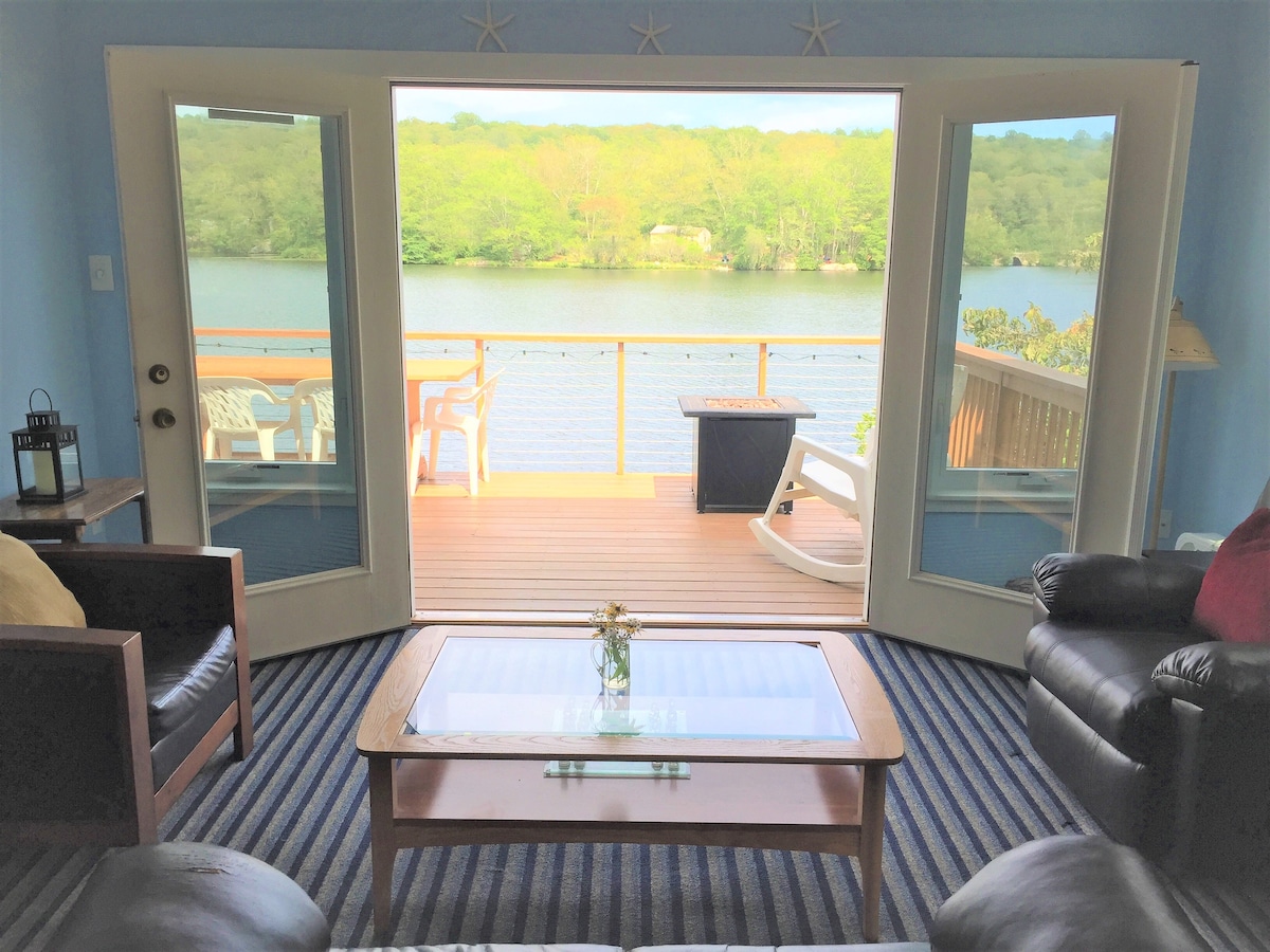 The living area features large French doors that open to a scenic view of the lake, inviting natural light. A coffee table sits center, adorned with a small vase of flowers. Comfortable seating includes two leather couches facing the lake, with striped area rugs underfoot.