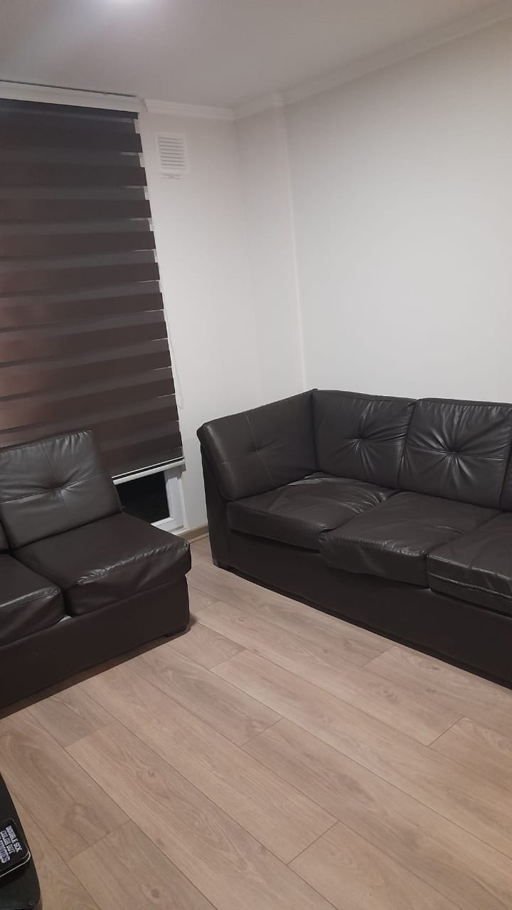 A cozy living area features a dark leather sectional sofa positioned against a neutral, light-colored wall. A matching armchair complements the seating arrangement. Natural light filters through contemporary blackout curtains, and warm wooden flooring enhances the inviting space.