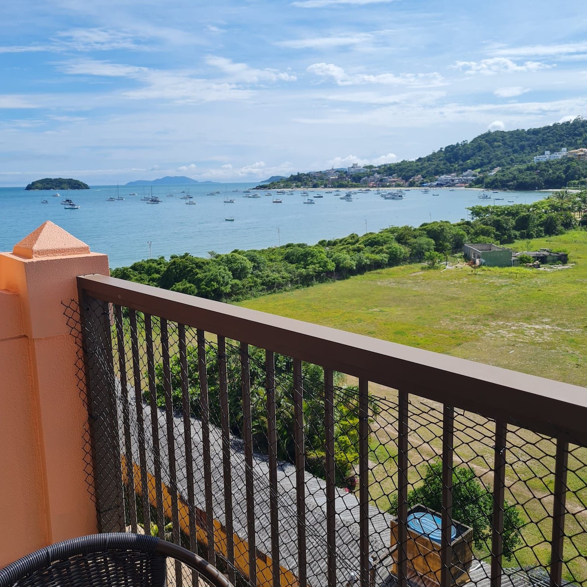 A balcony view is presented, showcasing a serene seascape with boats anchored in the distance. Lush greenery and hills are visible along the coastline under a partly cloudy sky, emphasizing the natural scenery surrounding the area.