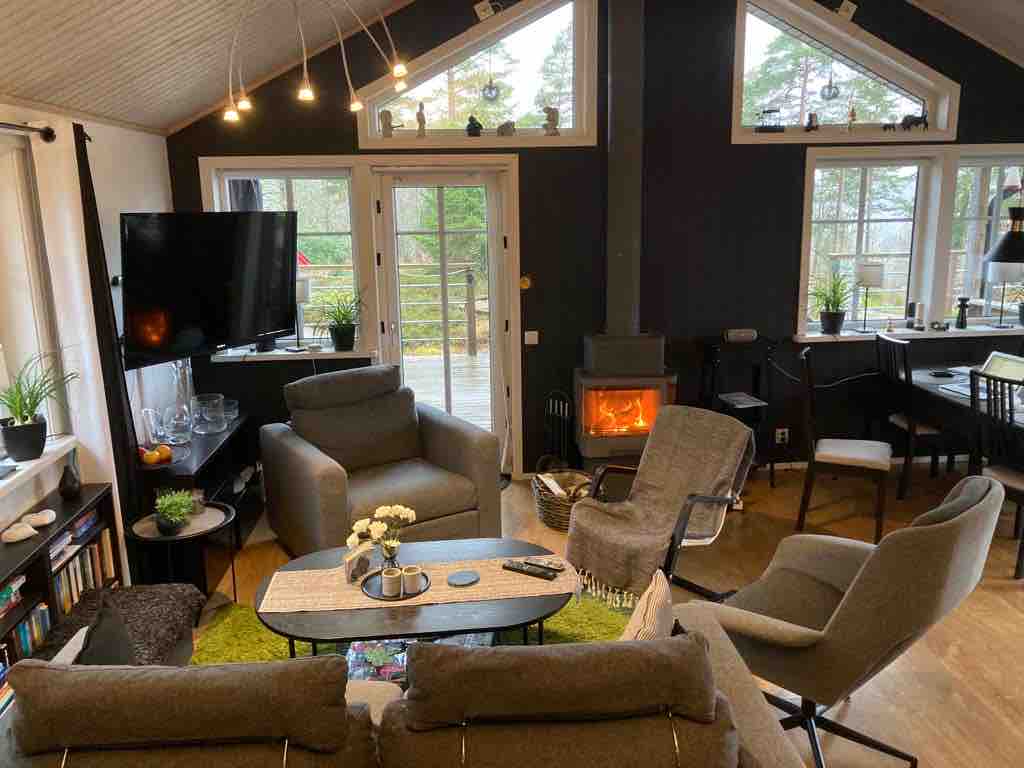 A cozy living area is highlighted by two large armchairs and a sofa surrounding a central coffee table. A television is mounted on the wall opposite the flickering flames of a fireplace, while large windows allow natural light and views of the outdoors.