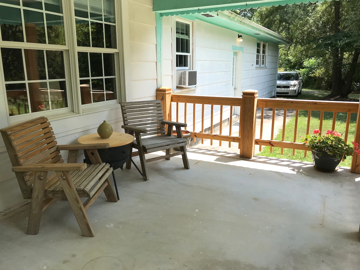 A covered patio area features two wooden chairs positioned around a small round table. A potted plant with colorful flowers is placed nearby, while a glimpse of the driveway and a parked vehicle can be seen in the background, surrounded by trees.