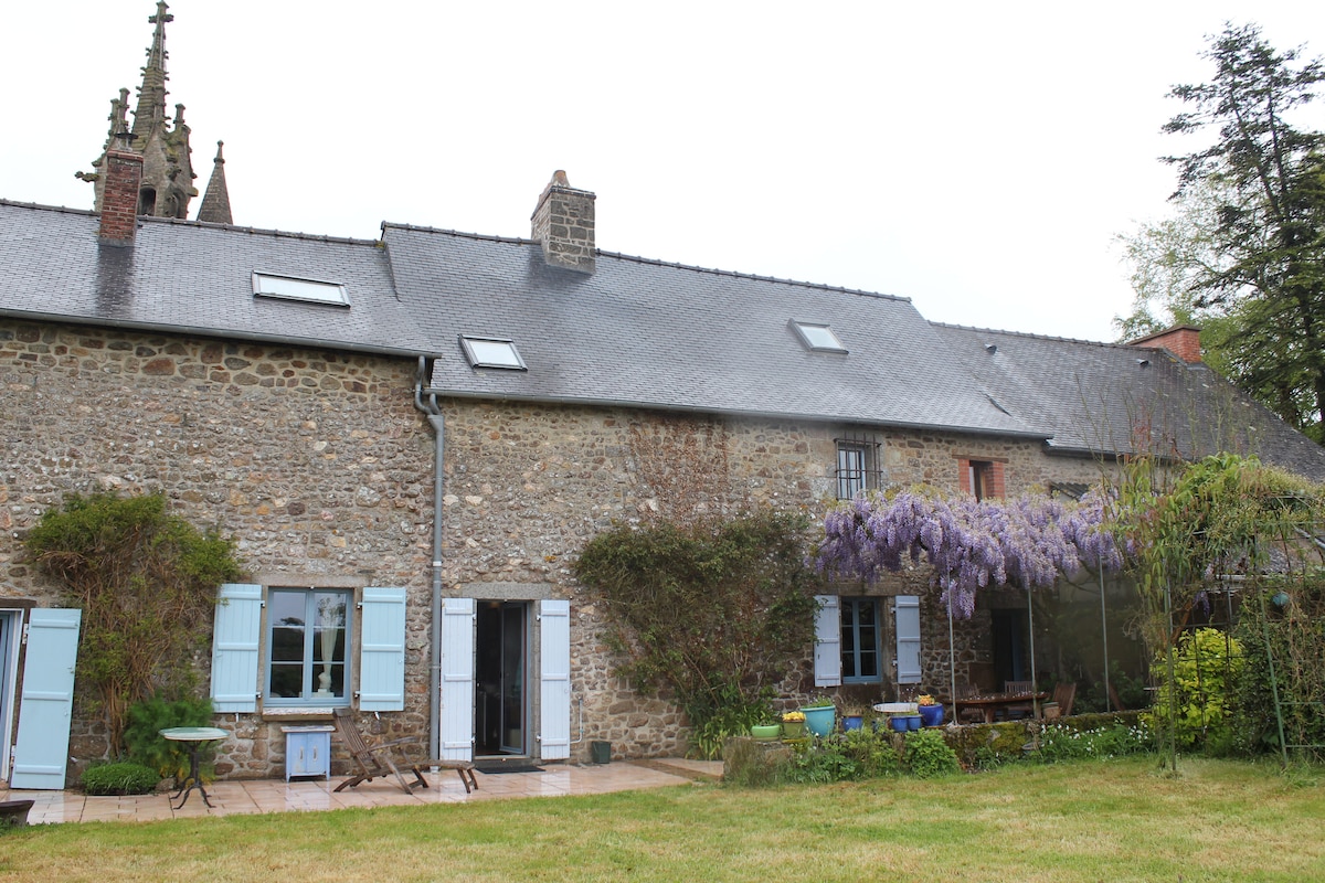 An old stone house is shown, featuring traditional blue shutters and a welcoming outdoor area. A wisteria vine is observed cascading over the exterior, complementing the greenery of the garden. Patio furniture is set up for outdoor relaxation, providing an inviting space for guests.