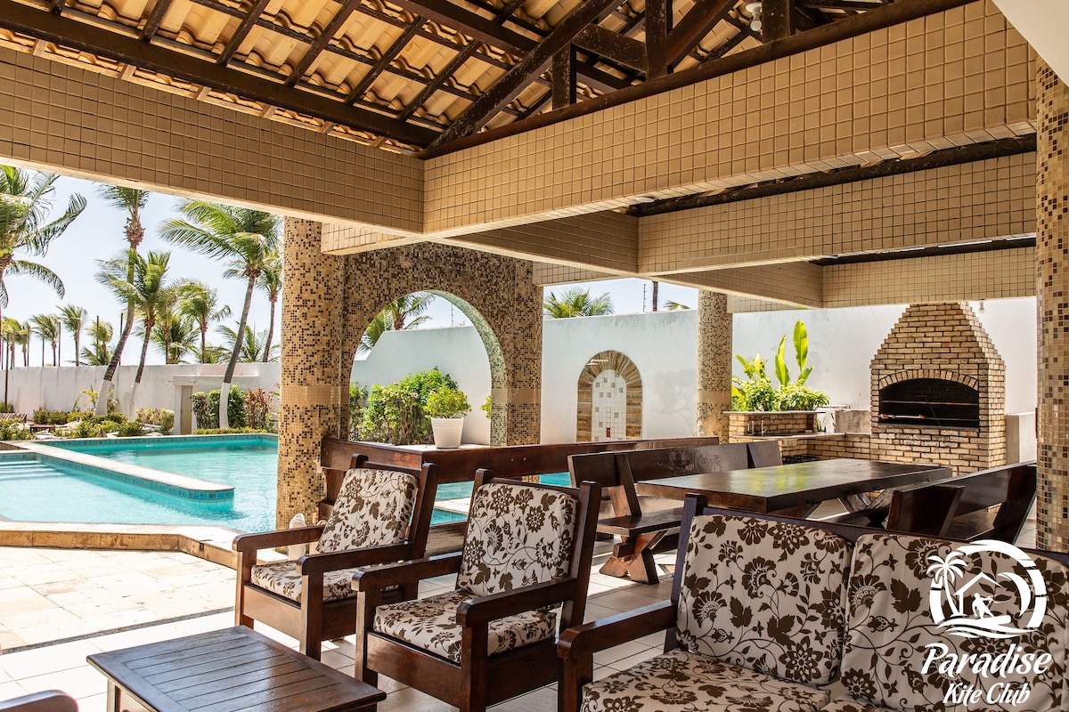A shaded outdoor seating area features wooden furniture with floral-patterned cushions. The view includes a pool and lush palm trees, contributing to a relaxed ambiance. A brick barbecue stands off to the side, framed by decorative arches and warm, natural lighting.
