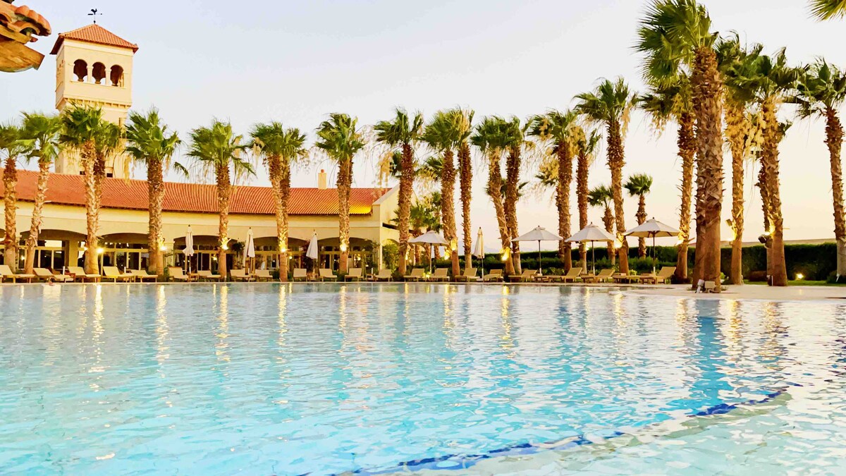 A spacious swimming pool reflects the palm trees and the resort's architecture. Lounge chairs line the poolside, and umbrellas provide shade. The setting is complemented by a building in the background with a red-tiled roof and a tower, presenting a relaxed atmosphere.