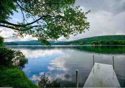 A calm lake scene is depicted, with still water reflecting the sky and surrounding greenery. A wooden dock extends into the water, framed by lush foliage and distant hills, creating a serene and inviting atmosphere for relaxation.