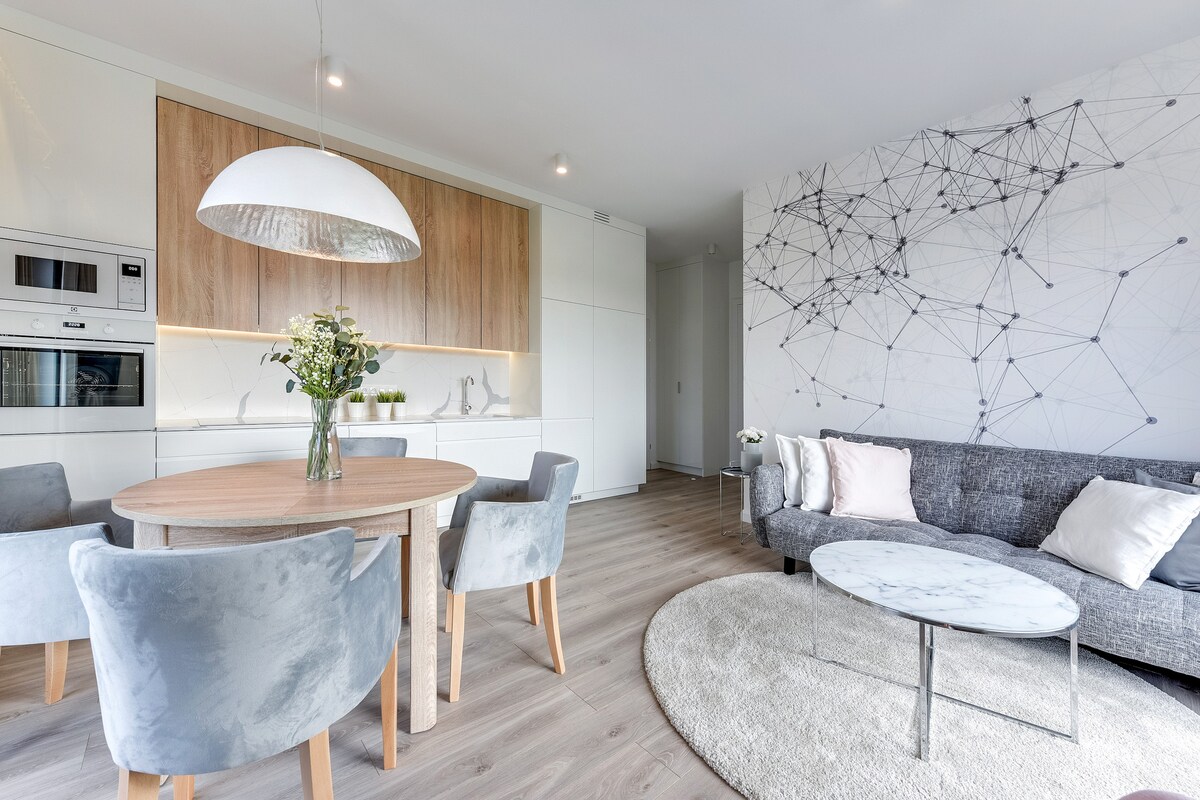 A modern living area is presented, featuring a round dining table surrounded by soft chairs. Light wood cabinetry and sleek appliances are visible in the background. A cozy gray sofa with decorative pillows is positioned next to a circular rug, creating an inviting space.