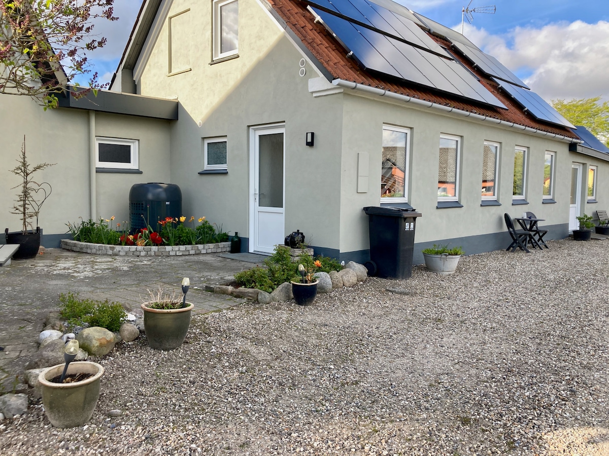 The exterior of a light-colored house is depicted, featuring a well-maintained garden with flower beds and decorative pots. Solar panels are installed on the roof. A gravel pathway leads to the entrance, flanked by a water tank and recycling bins. Two outdoor chairs are situated beside the house.