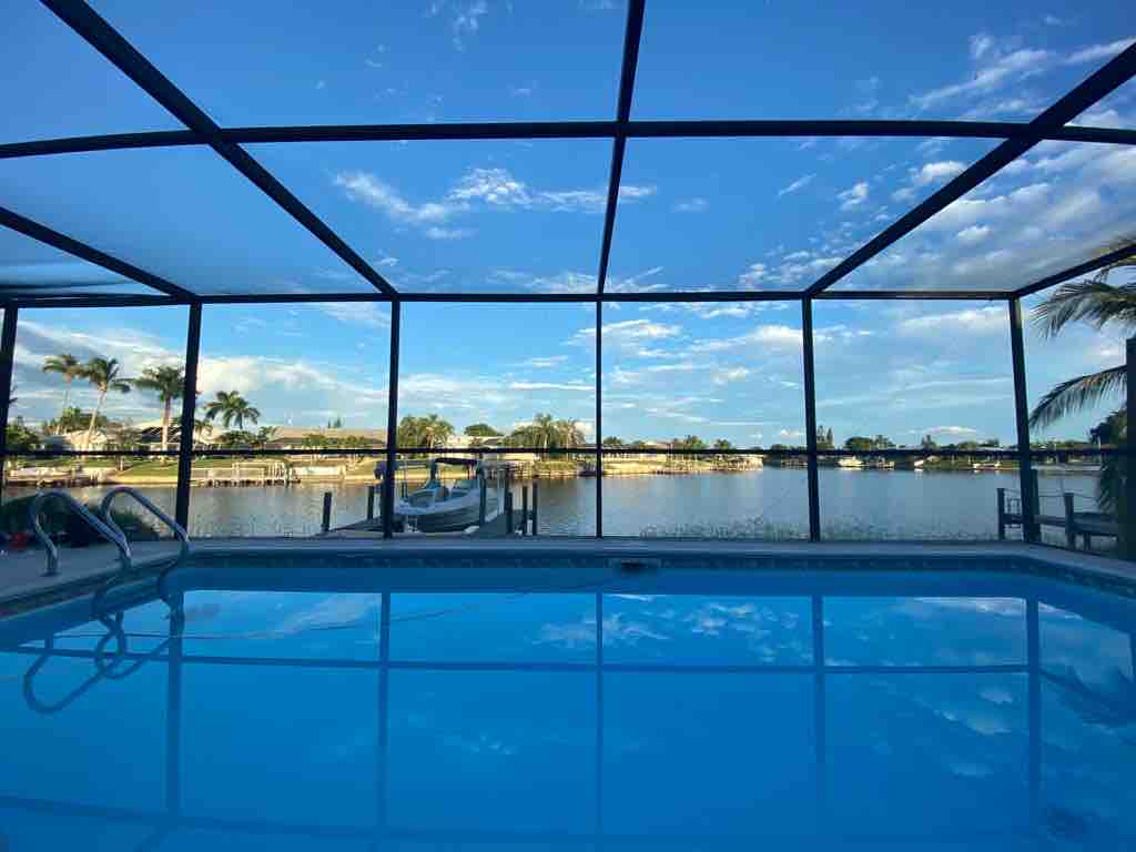 A serene view showcases a refreshing pool enclosed by a screened patio, with clear blue water reflecting the sky. The expansive glass panels reveal a wide canal and lush surroundings, offering a glimpse of nearby boats and waterfront homes.