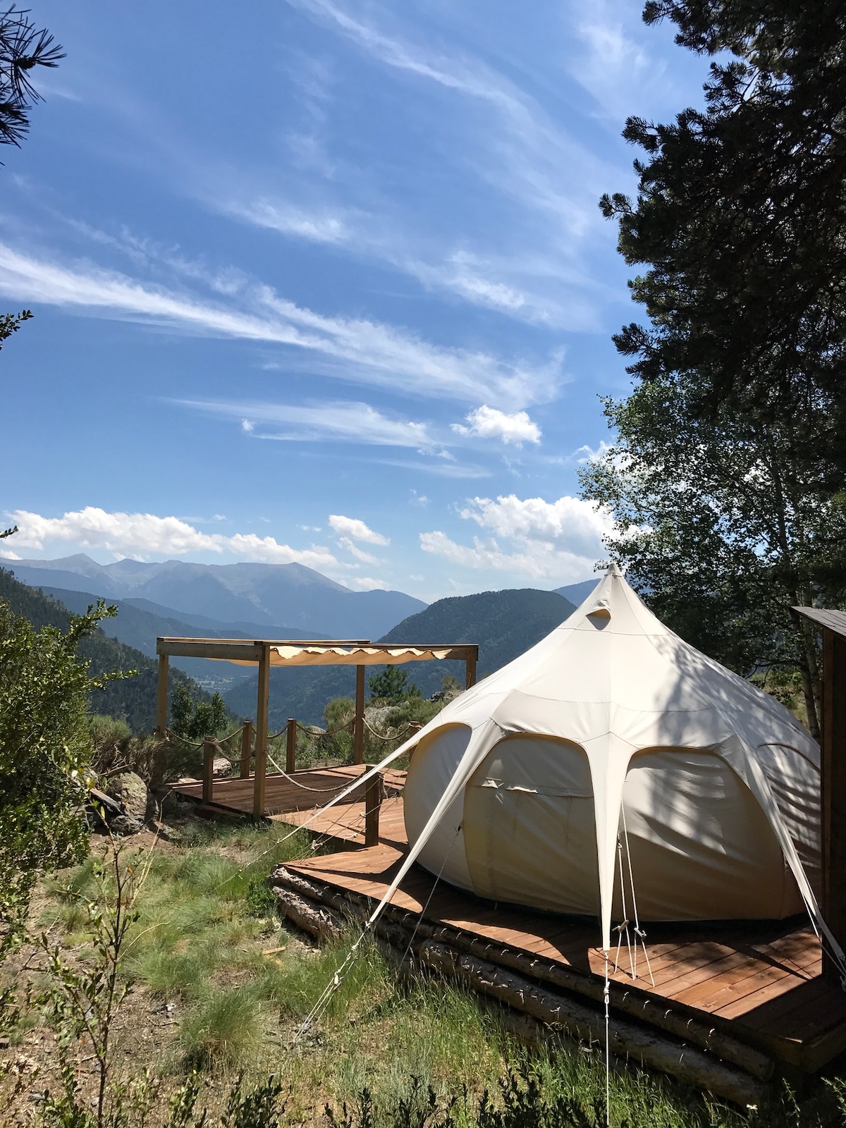 A spacious tent is set on a wooden deck, surrounded by lush greenery. The mountains are visible in the background under a clear blue sky. A shaded area with a wooden structure complements the outdoor setting, enhancing the natural ambiance.