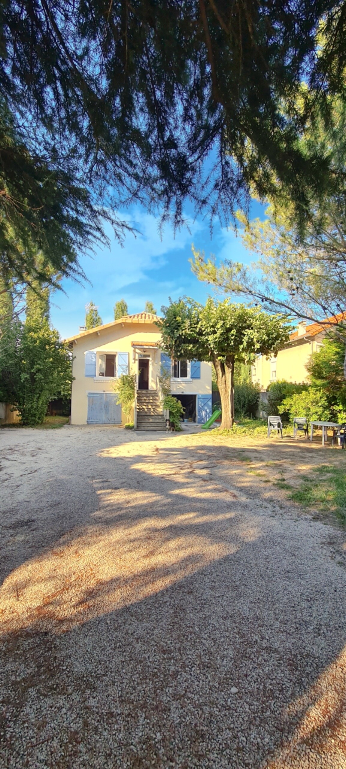 An exterior view of a two-story home surrounded by lush greenery. The gravel pathway leads to a charming entrance with blue shutters and a staircase. Sunlight filters through the trees, creating soft shadows on the ground.