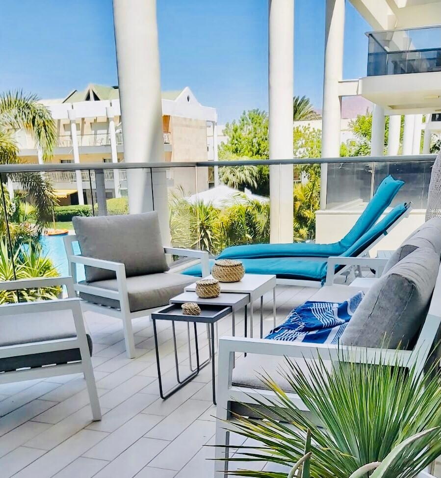 A spacious terrace is showcased, featuring comfortable seating arrangements including two armchairs and a chaise lounge. A small table holds decorative items. Lush greenery can be seen beyond the glass railing, contributing to the calm ambiance.