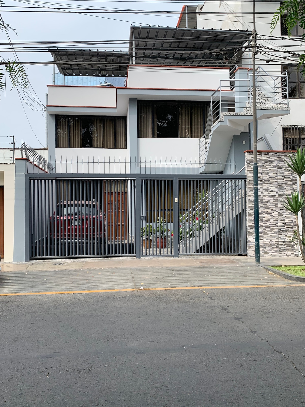 The exterior of the property is shown, featuring a multi-story structure with large windows and a balcony. A gated entrance leads to the driveway, where a car is parked. The building is surrounded by greenery, adding a touch of nature to the urban setting.