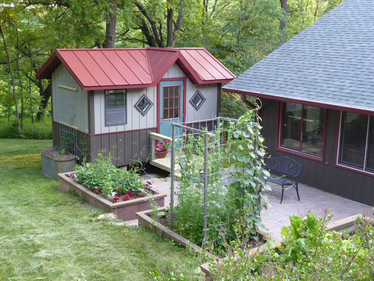 A charming tiny house with a red metal roof is nestled among lush greenery. Flowering gardens are visible in raised beds, offering a vibrant backdrop. The adjacent structure features dark siding and large windows, creating a cozy outdoor atmosphere within a natural setting.