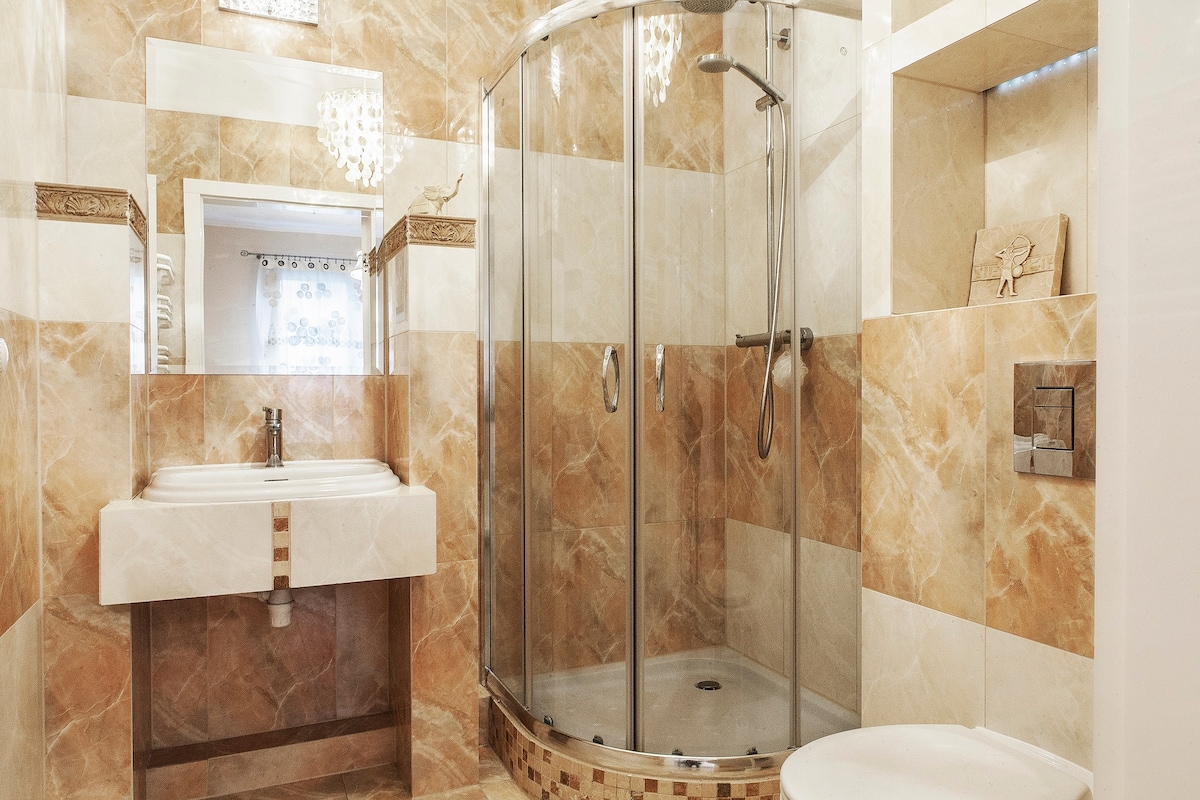 A modern bathroom features a glass-enclosed shower with a rainfall showerhead, while a counter-mounted sink is positioned above a minimalist vanity. Elegant marble tiles in warm hues line the walls, complemented by decorative accents, providing a clean and functional space.