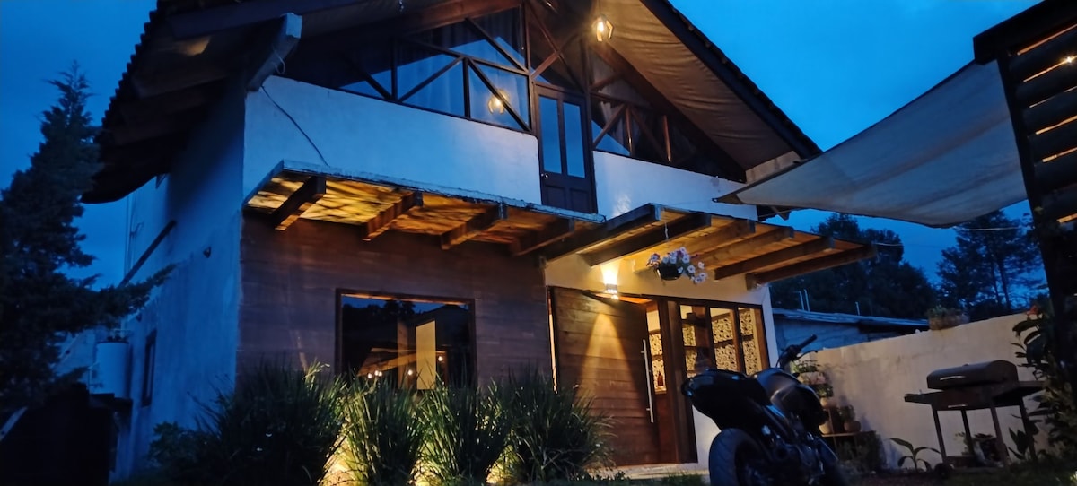 A two-story house is illuminated in soft light, showcasing wooden beams and a blend of rustic and modern elements. The exterior features large glass doors framed by lush greenery and a motorcycle parked nearby, contributing to a welcoming ambiance.