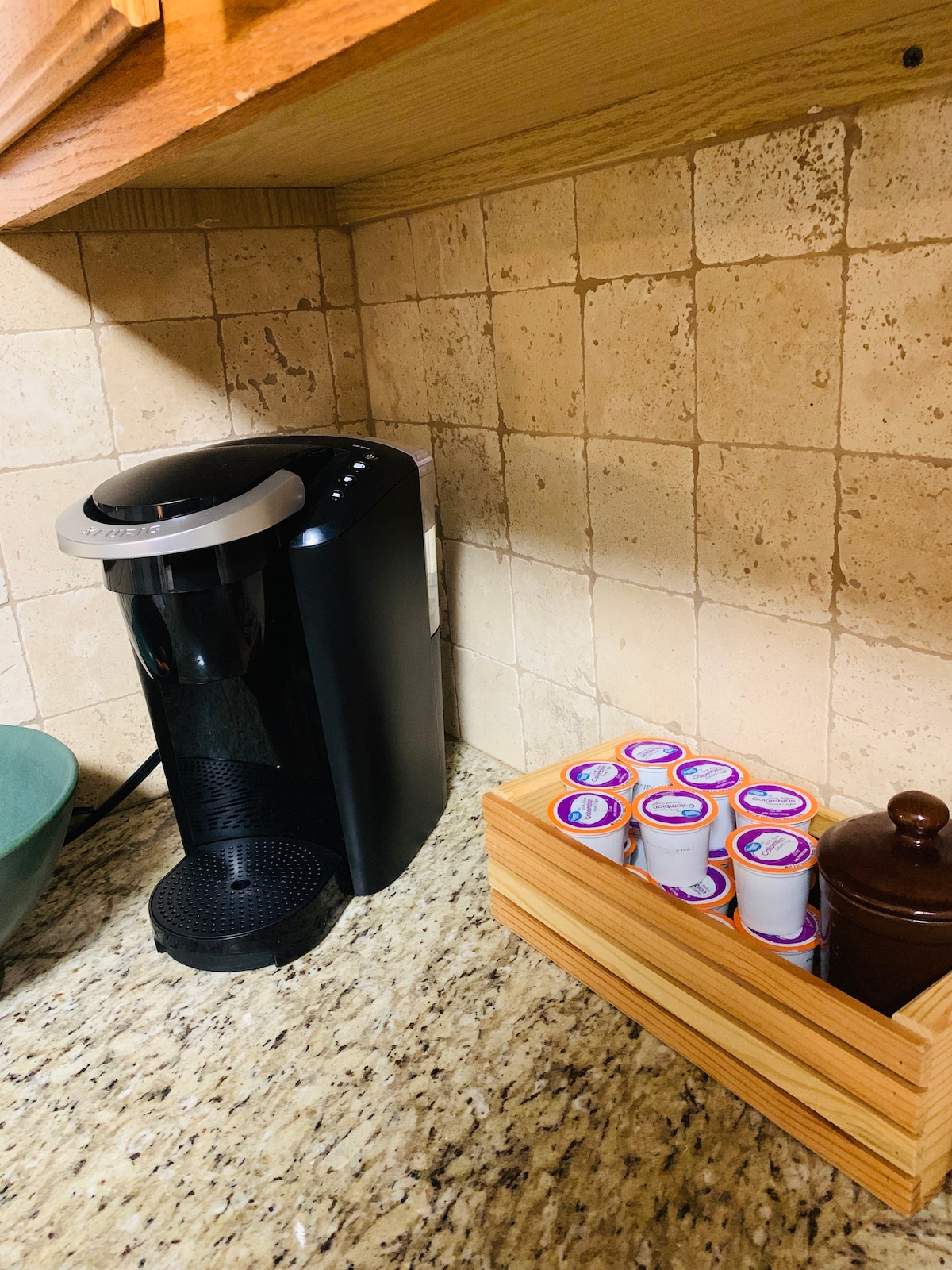A coffee maker is positioned on a granite countertop, accompanied by a wooden container holding an assortment of coffee pods. A small brown jar is also visible, adding to the organized appearance of the kitchen space.