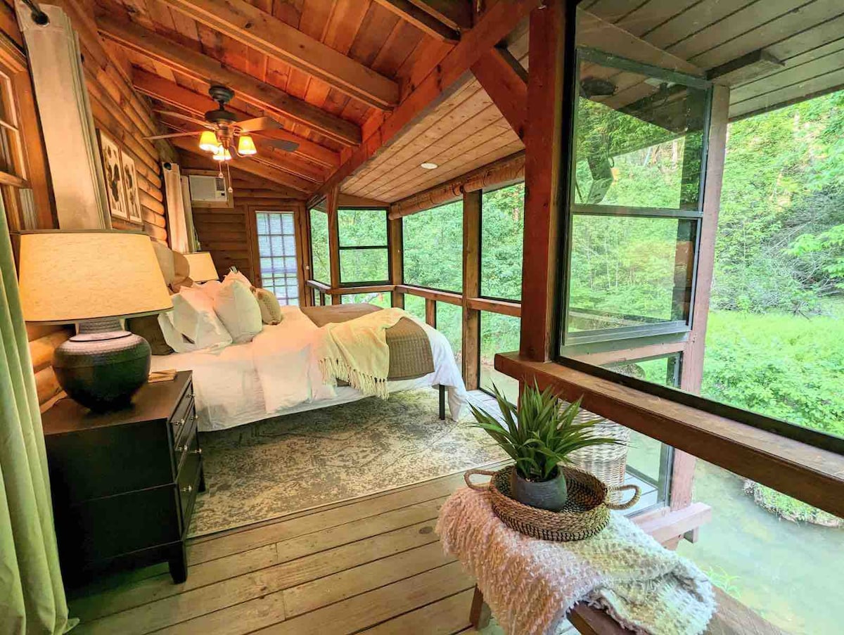 A cozy bedroom features wood-paneled walls and a vaulted ceiling, with large windows offering views of the surrounding greenery. A spacious bed is adorned with soft white linens, while a decorative lamp and plant add warmth to the space. A woven basket holds additional decor.