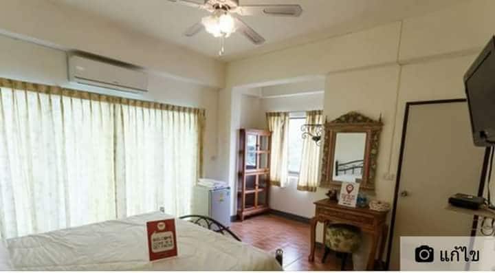 A standard room features large windows with sheer curtains allowing natural light to fill the space. A vintage-style wooden vanity with a mirror is situated beside a seating area. A comfortable bed is complemented by a ceiling fan, adding to the room's airy feel.