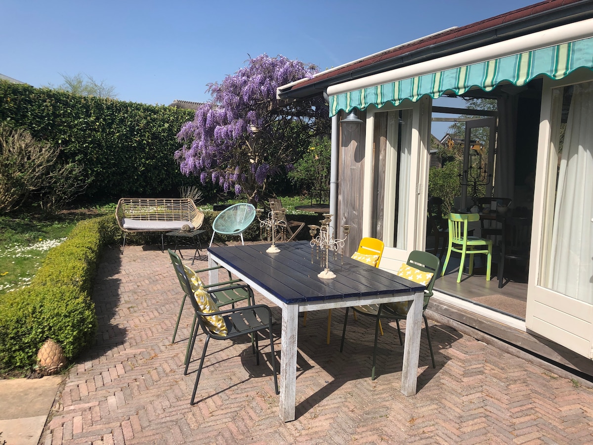 An outdoor dining area features a large table surrounded by six chairs in various colors. A flowering tree provides a natural backdrop, while the patio is paved with brick. Green hedges and an additional seating area contribute to a spacious, inviting garden environment.