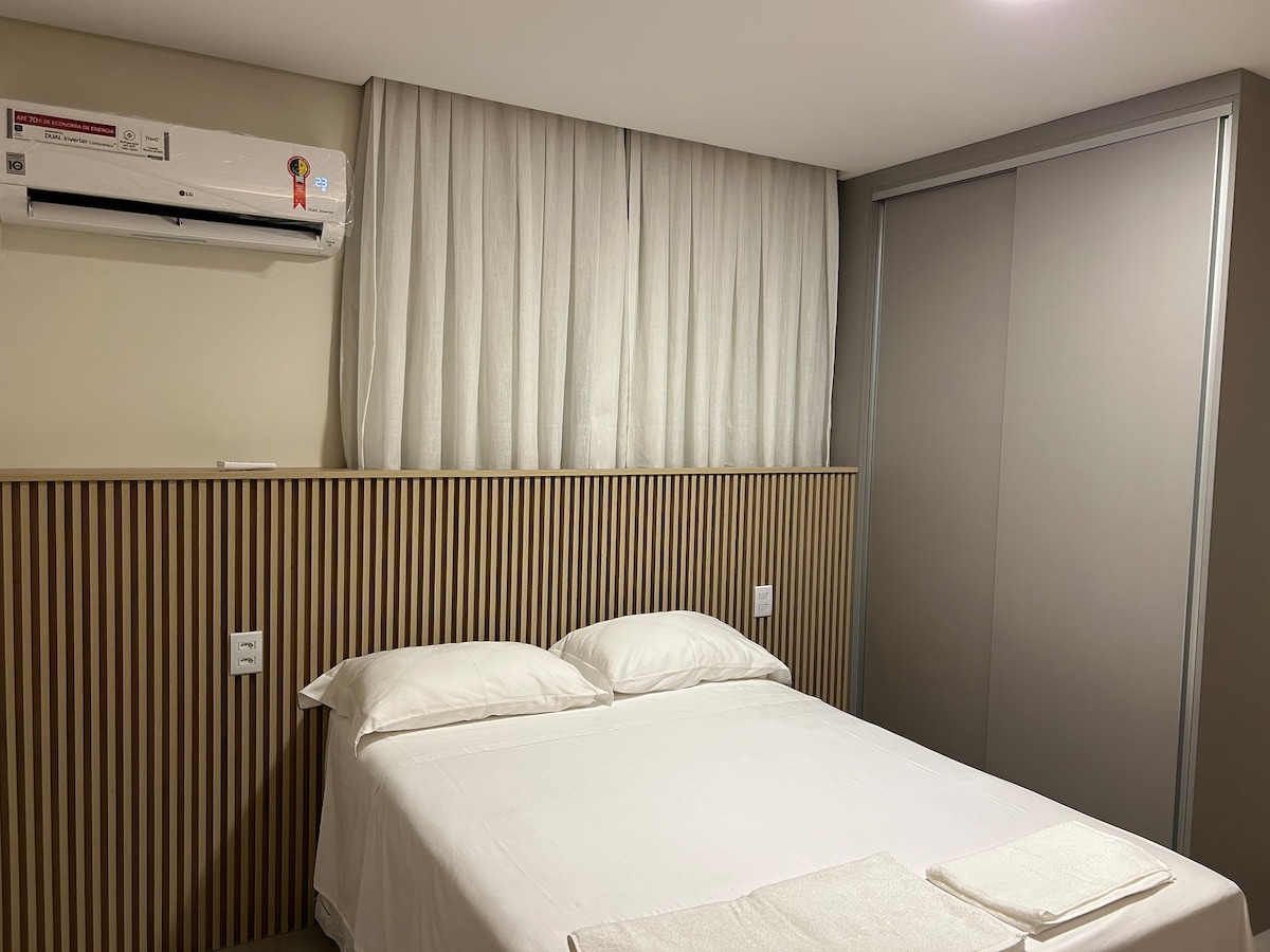 A clean bedroom features a spacious bed with white linens. Light-colored wood paneling enhances the headboard area, while sheer curtains cover a window, allowing soft light into the room. An air conditioning unit is mounted above, and a built-in closet stands nearby.