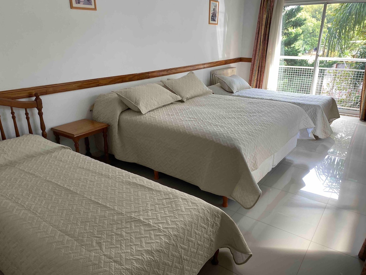 A spacious sleeping area features three neatly made beds with soft, neutral bedding. A wooden nightstand stands beside one of the beds. Large windows allow natural light to enter, while light curtains frame the view of greenery outside.
