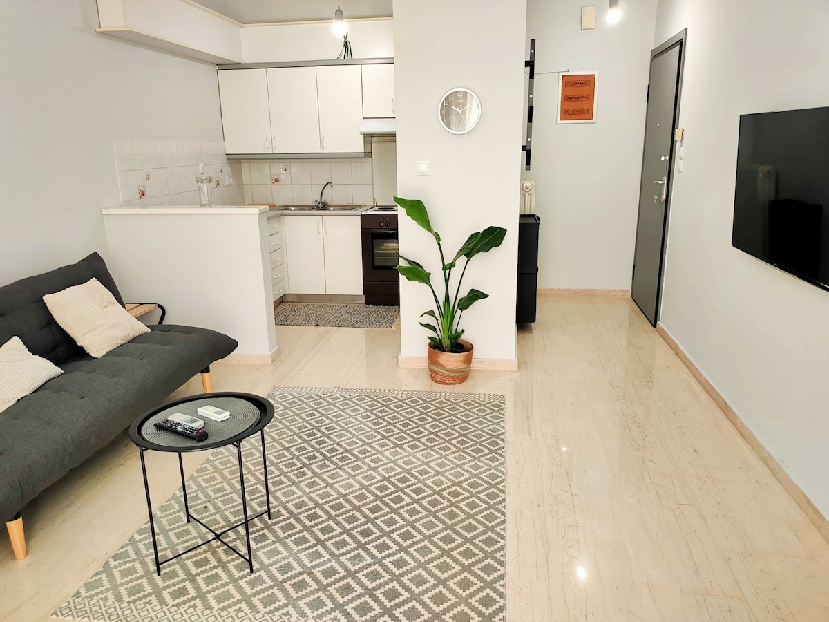 An open living space features a comfortable gray sofa and a small black coffee table. A kitchen area with white cabinets is visible in the background. A potted plant adds a touch of greenery, while a wall clock is mounted near the entrance.