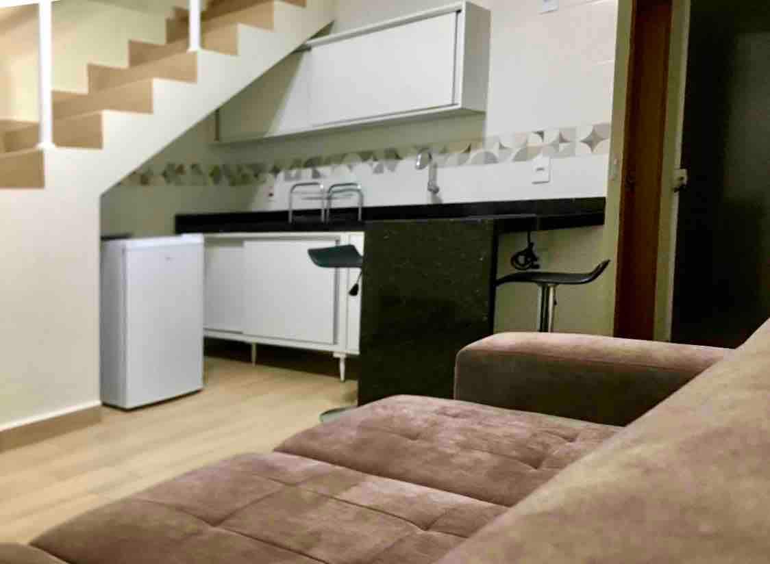 A compact kitchen area is visible, equipped with a white refrigerator, a sink, and a black countertop. A high chair is positioned near the counter. The space is enhanced by light wood flooring and modern cabinetry, with stairs leading to an upper level.