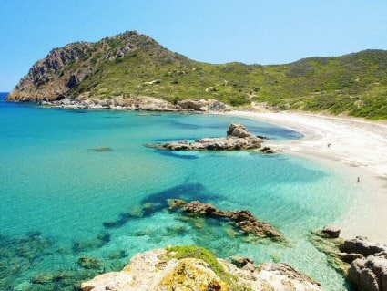 A serene beach scene presents clear turquoise waters lapping against a sandy shore. Rocky outcrops rise from the water, while gentle hills covered in greenery frame the landscape under a bright blue sky.
