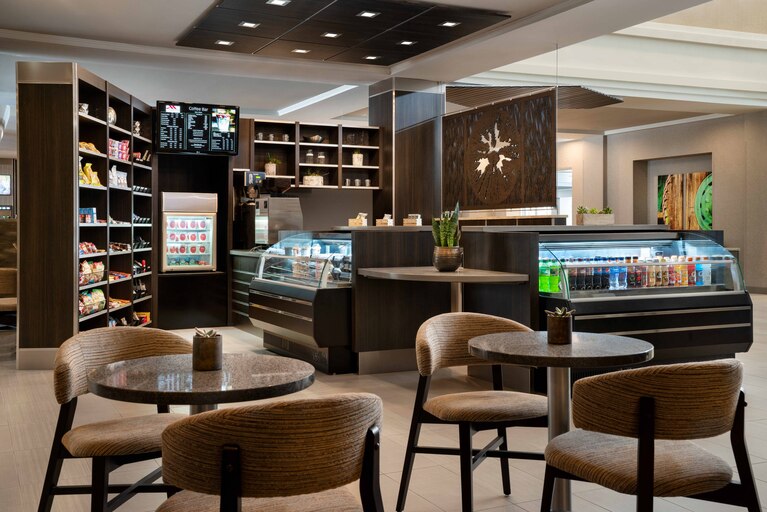 A modern café space features a sleek counter displaying a variety of food and beverage options. Plush seating is arranged around small tables, with shelves stocked with snacks and drinks visible in the background. Natural light enhances the inviting atmosphere of the space.