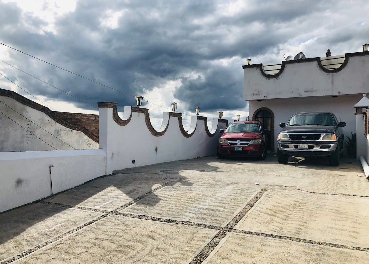 A spacious parking area is seen, accommodating two vehicles. The surface is paved, with patterned details visible. The surrounding walls are painted white, featuring decorative elements at the top. Overhead, clouds gather, creating a dramatic sky, while soft lighting fixtures line the perimeter.