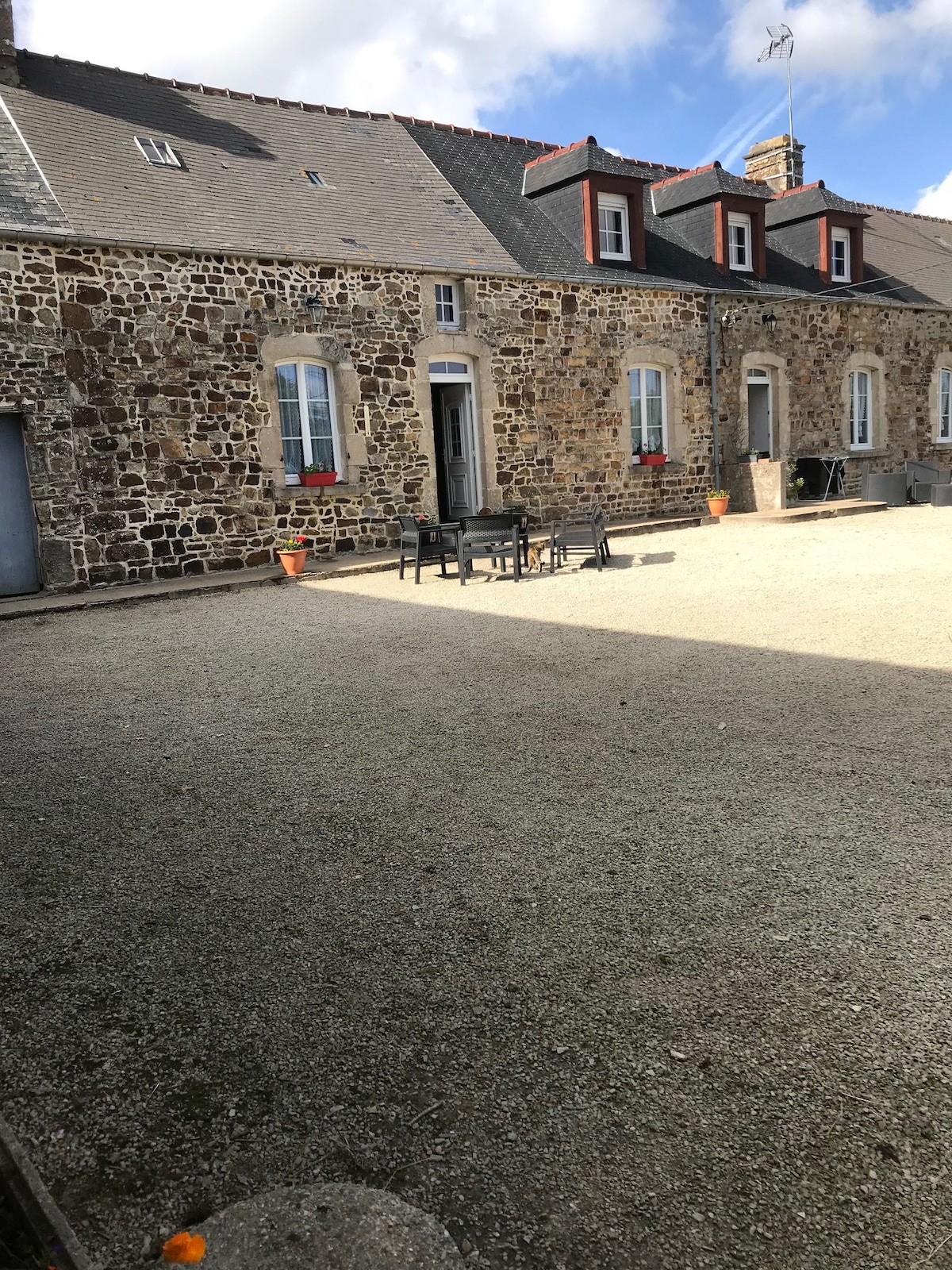 An outdoor courtyard is shown, featuring a stone building with several windows adorned with flower pots. A set of outdoor furniture is arranged on the gravel surface, providing a space for relaxation. Bright sunlight creates a natural contrast with shaded areas across the courtyard.