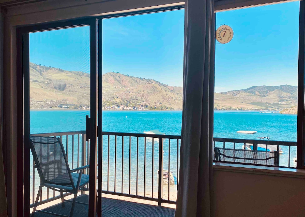 A stunning lake view is captured through the sliding glass door, showcasing clear blue water and distant hills. A single chair is positioned on the balcony, allowing for relaxation while enjoying the picturesque surroundings.