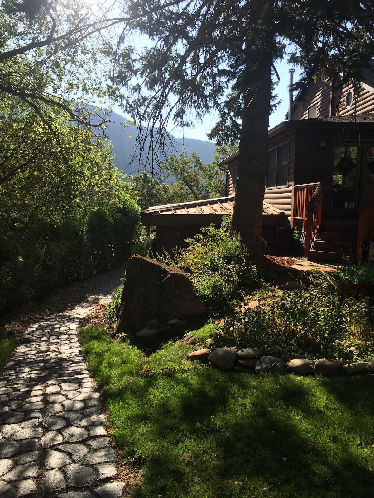 A stone pathway meanders through lush green grass and vibrant foliage, leading to a cabin structure with wooden elements. Sunlight filters through trees, casting a warm glow on the exterior, while distant mountains are visible beyond the foliage.