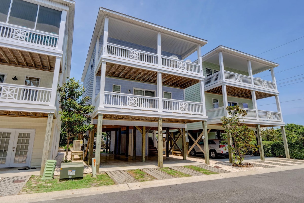 The exterior of the beach house is visible, showcasing three levels with spacious balconies adorned with white railings. The ground level includes covered parking beneath the house. Nearby lush trees and a paved driveway contribute to the home's inviting curb appeal.