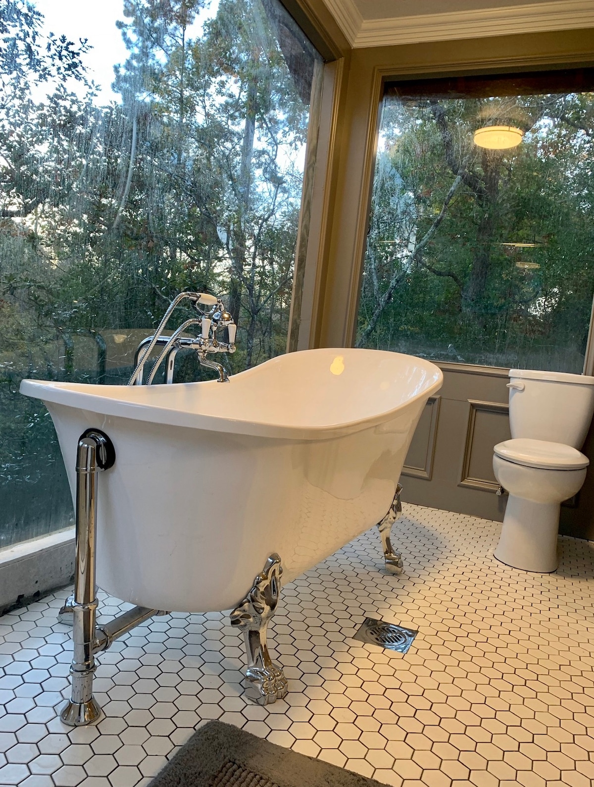 A freestanding white bathtub with chrome fixtures is positioned in an elegantly designed bathroom. Large windows provide a view of the surrounding trees, enhancing the natural light. The floor is covered in hexagonal tiles, and a white toilet is situated nearby.