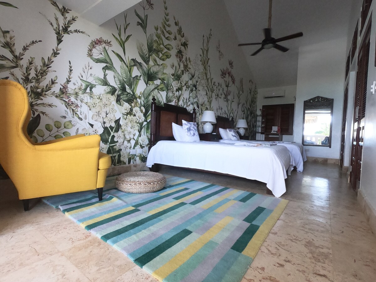 A spacious guest room features a dramatic botanical mural and two beds with crisp white linens. A comfortable yellow armchair adds a pop of color, while a patterned area rug softens the floor. Natural light filters through a nearby window, enhancing the serene atmosphere.