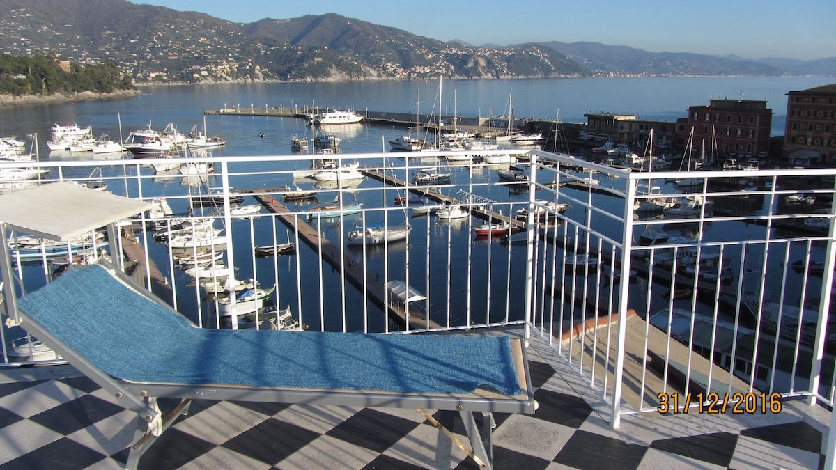 A terrace features a lounge chair overlooking the marina, showcasing a panoramic view of the water and surrounding hills. Numerous boats are docked in the harbor, with the coastline visible in the background under clear skies.