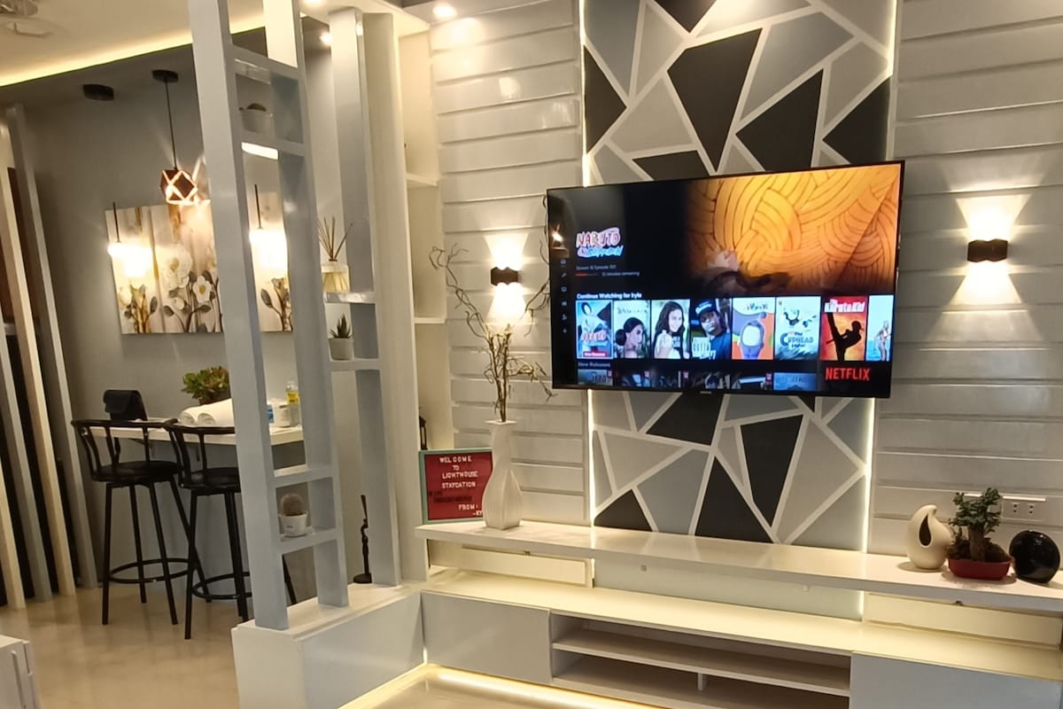 A modern living area is displayed with a large flat-screen TV mounted on a textured wall. A minimalist entertainment unit is positioned beneath the TV. Bar stools and a dining table are visible in the background, complementing the fresh and open ambiance of the space.