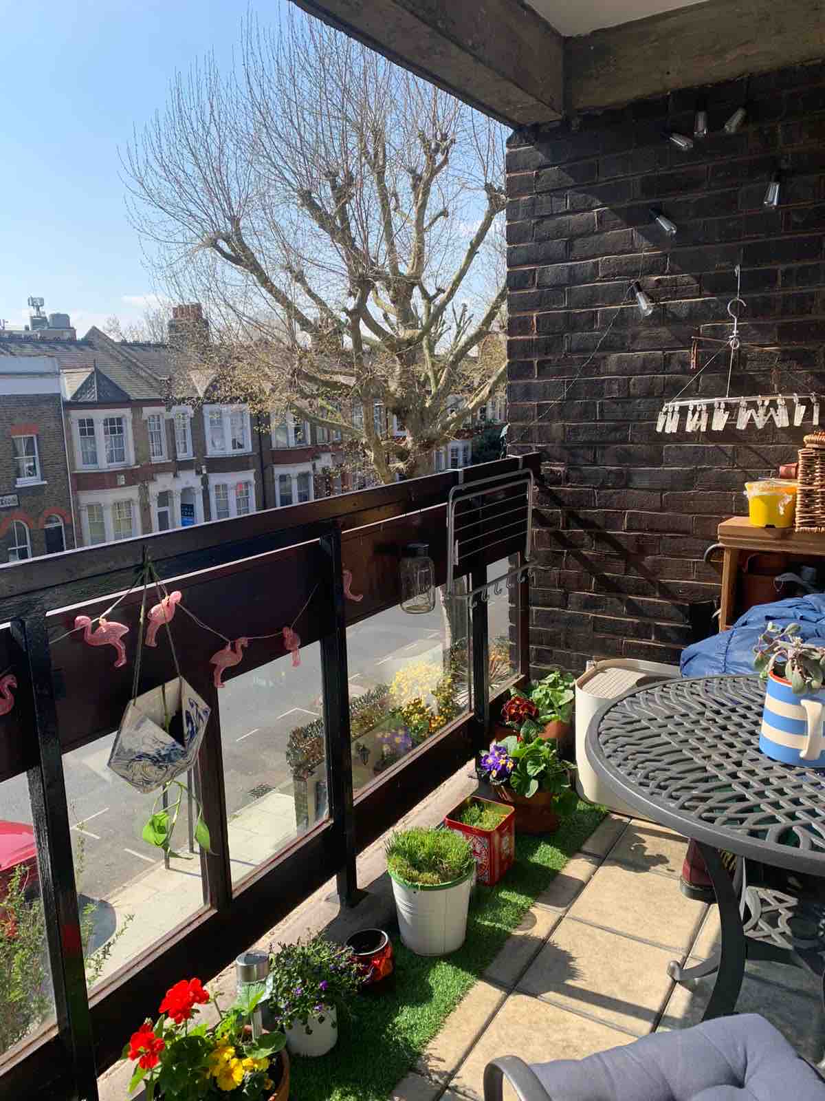 A sunny balcony can be seen, featuring a round table with chairs and adorned with colorful flower pots. Decorative flamingos hang along the railing, while the street view displays charming residential buildings in the background. A tree with bare branches is visible against a clear sky.