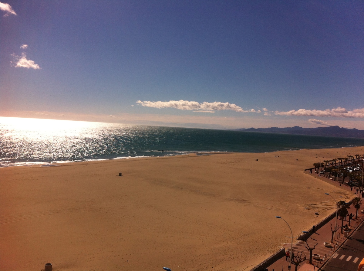 A vast sandy beach stretches along the shoreline, meeting the glistening sea under a clear blue sky. Gentle waves can be seen lapping at the edge of the sand, with distant mountains visible on the horizon.
