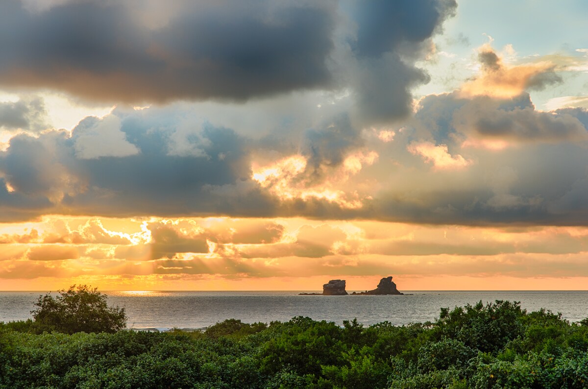 The image captures a scenic view of the ocean at sunset, with clouds reflecting warm hues of orange and yellow. Two rock formations can be seen in the distance, silhouetted against the vibrant sky. Lush greenery is featured in the foreground, enhancing the natural beauty of the scene.