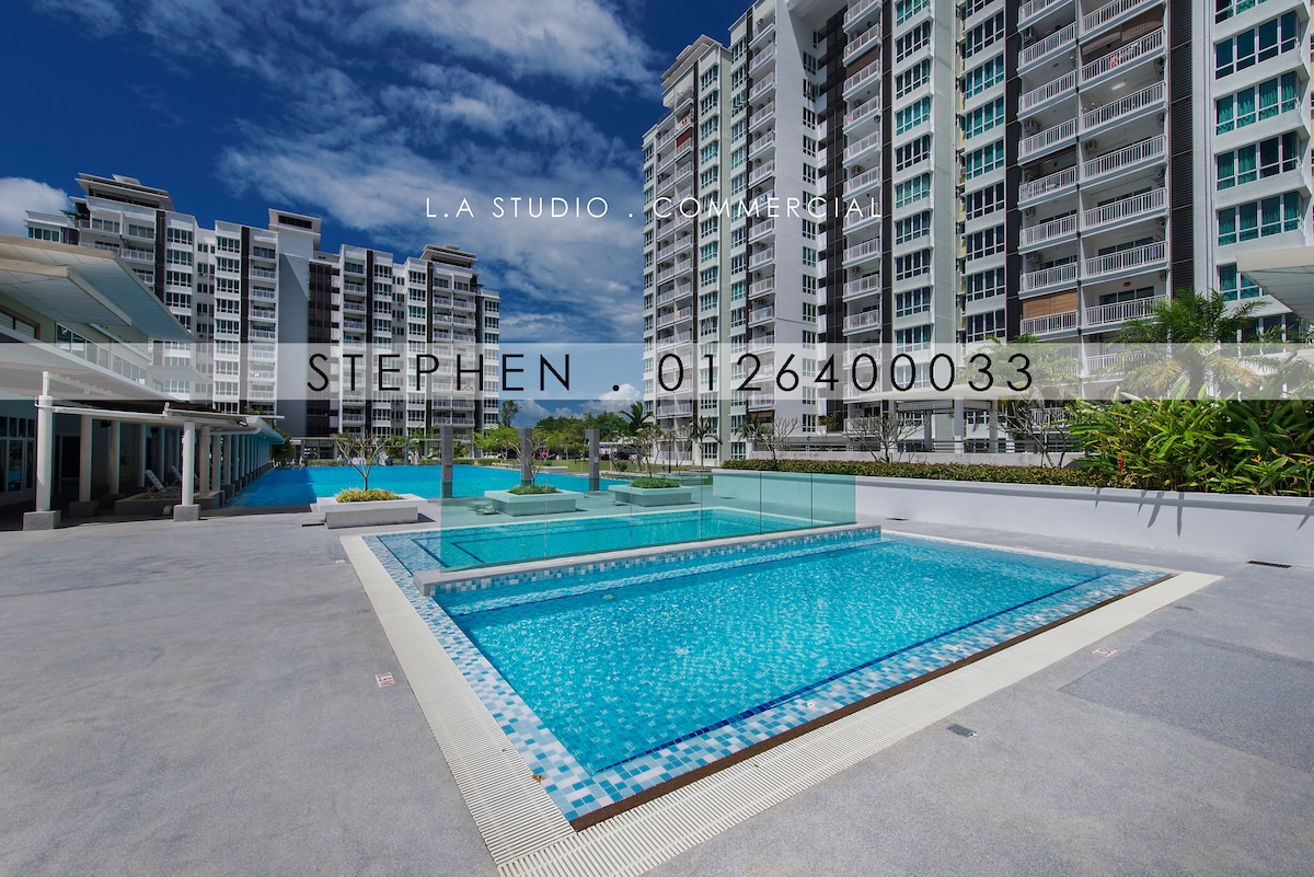 A well-maintained swimming pool area is visible, surrounded by lush greenery and modern buildings. Sun loungers are placed along the poolside, providing spots for relaxation. The pool features a clear blue surface, reflecting the sky above, creating an inviting environment for guests.