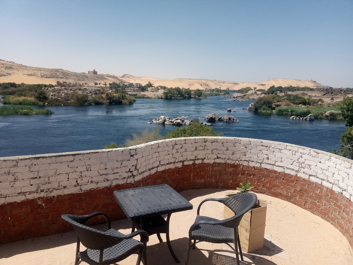 A terrace area presents a view of a river bordered by lush vegetation and sandy dunes in the background. Two wicker chairs are placed around a small black table, inviting relaxation. The serene water reflects the clear blue sky above.
